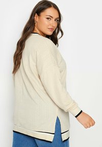 Top beige a maniche lunghe con motivo testurizzato, bordi neri e crema, spacchi laterali, vestibilità comoda e polsini a costine. Abbinato a un paio di jeans azzurri chiari.