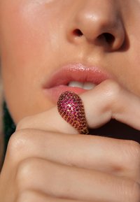 Bague en or en forme de goutte, incrustée de pierres précieuses roses, tenue contre une main à la peau douce et à la couleur des lèvres naturelle.