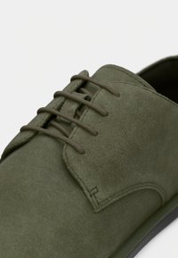 Πράσινο σουέτ παπούτσι με ομαλή υφή, διαθέτει πέντε τρύπες για κορδόνια και στρογγυλεμένη μύτη. Η σόλα είναι σκούρα και ελαφρώς αντιθετική.