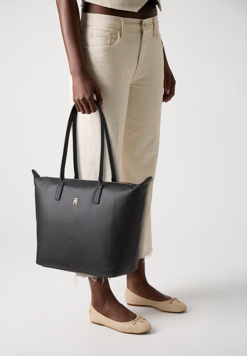 Sac fourre-tout noir avec double poignée, texture lisse, logo en métal doré sur le devant, forme rectangulaire, associé à un pantalon beige et des ballerines.