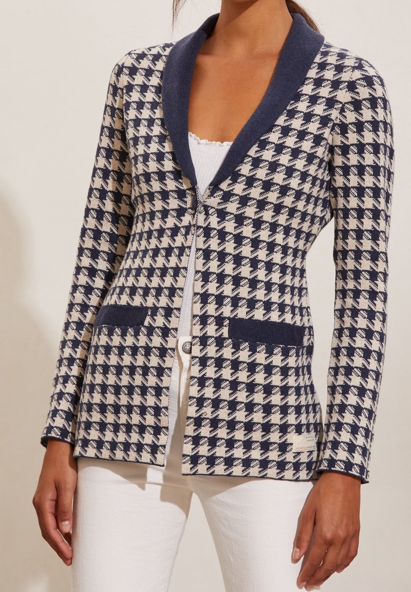 Femme portant un blazer à motif pied-de-poule bleu marine et beige avec un col bleu marine foncé et des détails de poches, par-dessus un haut blanc et un pantalon blanc.