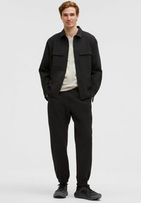 Mustmustükk must kaksiosaline riietus, mis sisaldab kraega särki nööbitud taskutega ja kitsenevaid pükse. Tekstureeritud kangas ja sportlikud jalanõud täiendavad välimust.