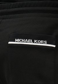 Poche en tissu noir avec une ligne horizontale blanche et "MICHAEL KORS" brodé en blanc au-dessus de la ligne.