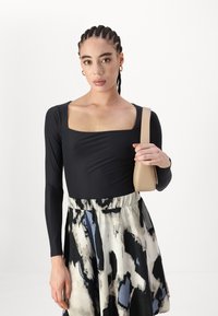 Musta pitkähihainen bodysuit, jossa on neliön muotoinen pääntie, yhdistettynä kuvioituun silkkiin viittavaan hameeseen, jossa on sinisiä ja mustia abstrakteja kuvioita. Pitelee vaaleanbeigeä käsilaukku.