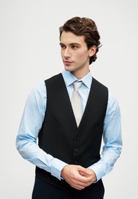 Junger Mann mit welligem braunem Haar, der ein hellblaues Hemd, eine cremefarbene Krawatte und eine schwarze, knöpfbare Weste trägt, schaut zur Seite mit gefalteten Händen.