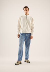 Balts fleeces pulovers ar pusīsiju rāvējslēdzēju, zilas džinsa bikses un daudzkrāsaini sporta apavi ar dzelteniem akcentiem un teksturētu zoli.