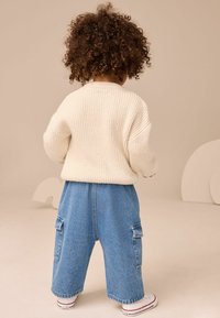 Maglione lavorato a maglia cremosa e pantaloni cargo in denim blu con tasche, abbinati a sneakers bianche con accenti rossi.