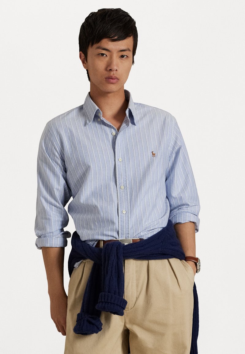 Lichtblauw gestreept button-up shirt van katoen, met lange mouwen en opgerolde manchetten, gecombineerd met beige geplooide broek en een marineblauw sweater geknoopt om de taille.