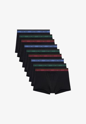 Hombre sin camiseta, de pie descalzo, vistiendo calzoncillos tipo bóxer negros con cintura verde sobre un fondo blanco liso.