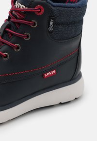 Botte imperméable Levi's noire et bleue avec coutures rouges, lacets rouges et noirs, et semelle blanche sur fond clair.