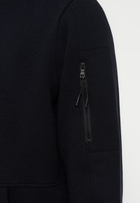 Μανίκι μαύρου μπουφάν με κάθετη τσέπη με φερμουάρ, που διαθέτει δύο τσεπάκια με φερμουάρ στο πάνω μέρος του βραχίονα, με ουδέτερο φόντο.