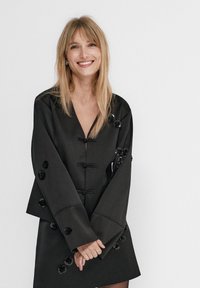 Robe noire à manches longues, avec un décolleté en V profond et des accents floraux décoratifs. Tissu lisse avec une silhouette structurée.