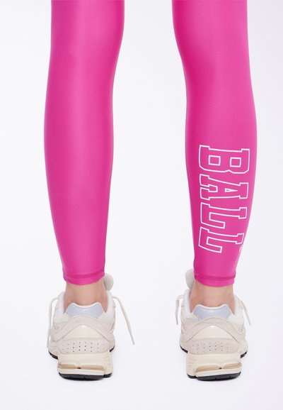 Prilagođene ružičaste tajice s bijelim natpisom "BALL" na donjem dijelu noge, uparene s svijetlim sportskim cipelama s suptilnim dizajnerskim detaljima.