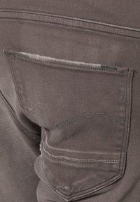 Grå denimtyg som visar en bakficka med subtil sömnad och en liten metallisk varumärkesetikett, med en mjuk textur och en bleknad utseende.