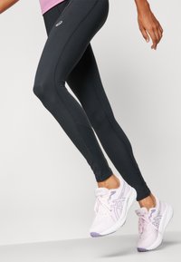 Zwarte sportleggingen met een slim fit en naaddetails, gecombineerd met lichtroze hardloopschoenen met witte zolen en paarse accenten.