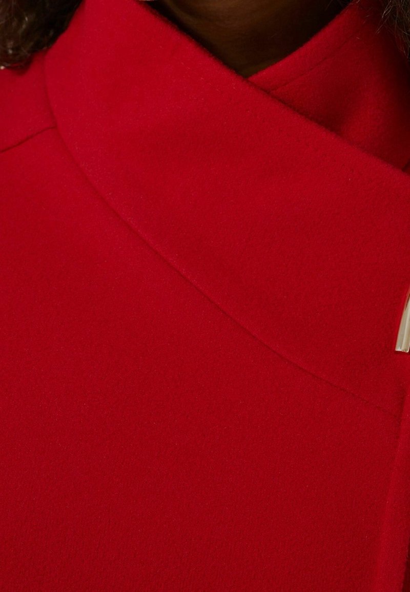 Gros plan d'un manteau en laine rouge avec un col haut superposé et une petite boucle métallique sur le côté droit.