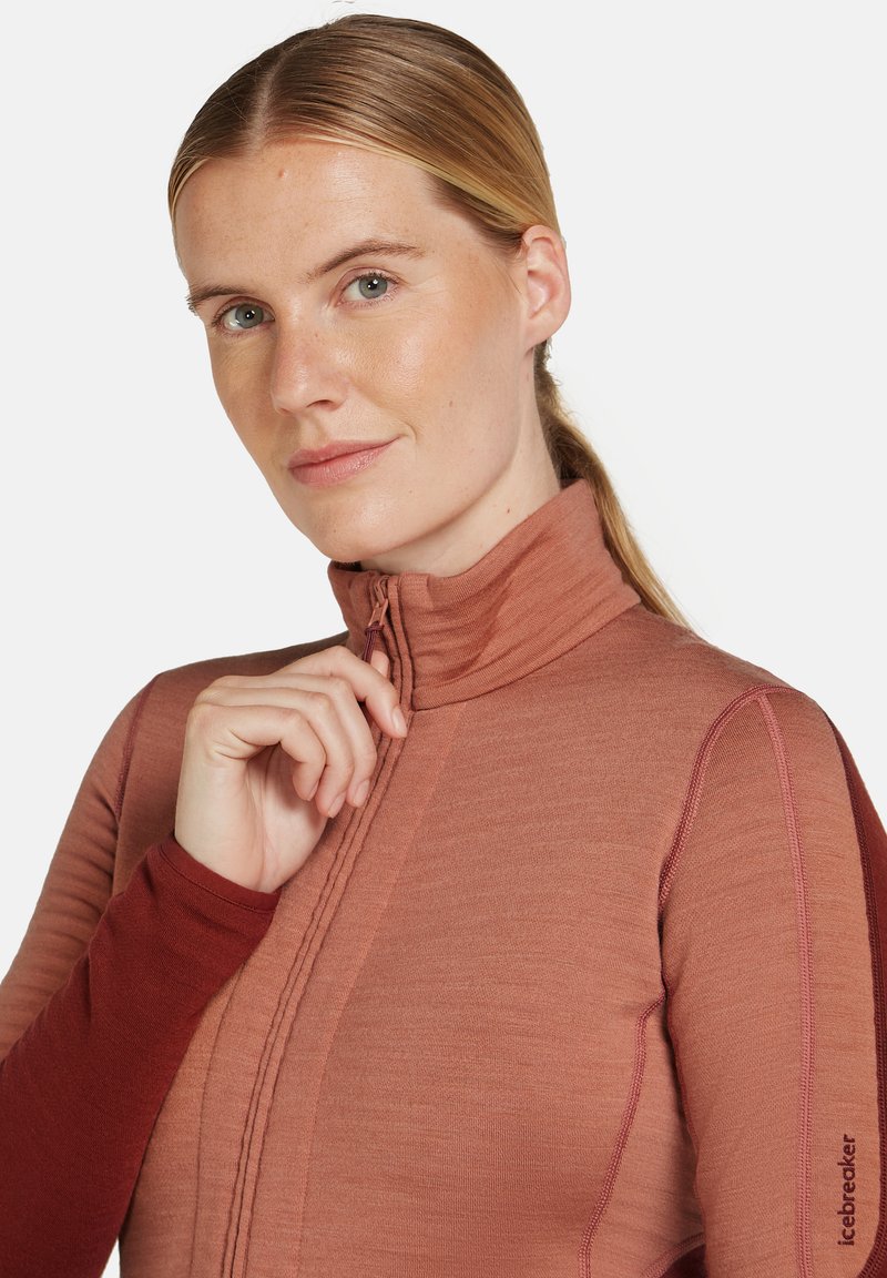 Langärmliges Zip-Top in Rost- und Burgundertönen, mit hohem Kragen, Nahtdetails und Logoakzent am Ärmel.