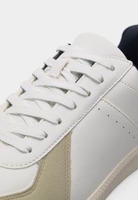 Λευκό αθλητικό παπούτσι με λευκά κορδόνια, μπεζ λεπτομέρεια από σουέτ κοντά στα δάχτυλα και ορατά ράμματα, απεικονίζεται σε κοντινή λήψη.