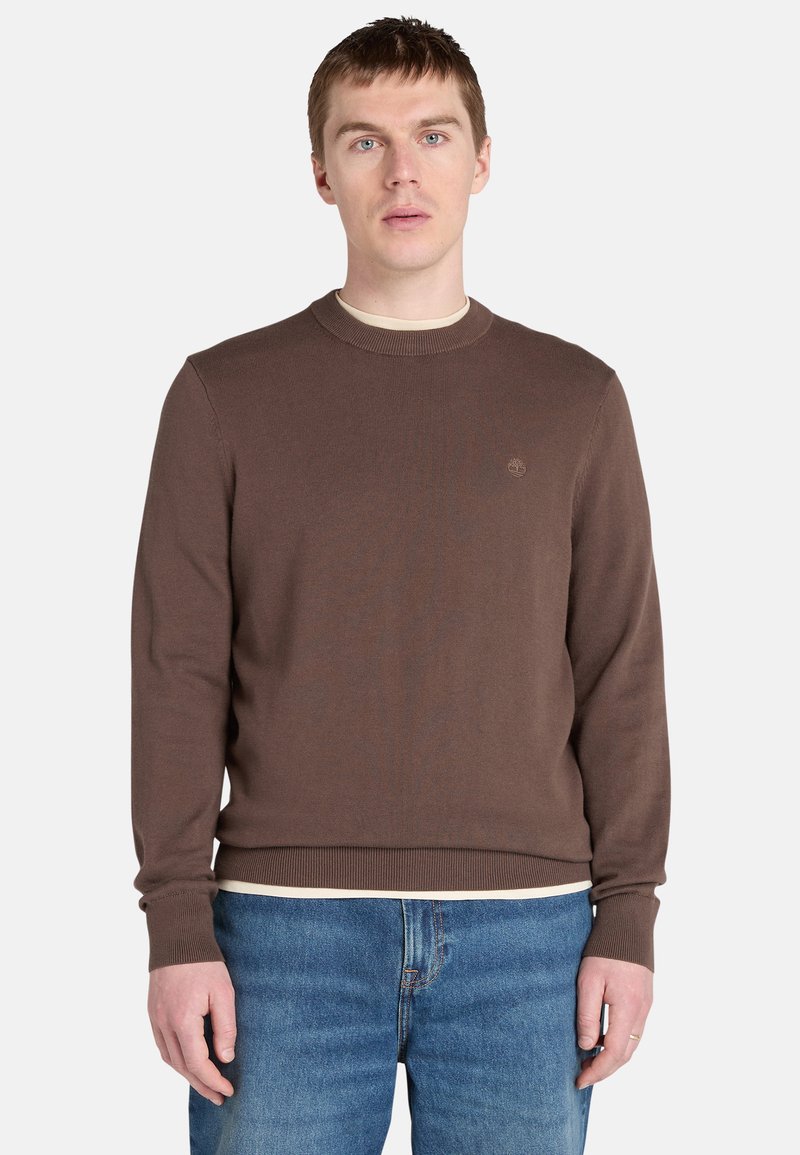 Brun stickad tröja med rund halsringning, ribbade ärmslut och nederkant, med en liten logotyp på bröstet. Bärs över en ljus skjorta.