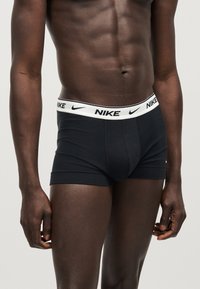 Γυμνασμένος ανδρικός κορμός που φοράει μαύρα μπόξερ Nike με άσπρη μέση που φέρει μαύρα λογότυπα Nike σε απλό ανοιχτό φόντο.