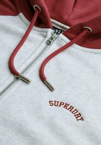 Sudadera con cremallera gris y capucha roja con cuerdas. Presenta herrajes de cremallera plateados y "SUPERDRY" bordado en rojo en el pecho.