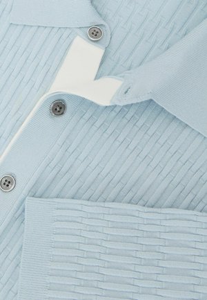 Világoskék bordázott kardigán ezüst gombokkal és fehér béléssel, a szövet textúrája jól látható közeli képen.