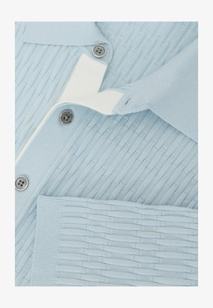 Világoskék bordázott kardigán ezüst gombokkal és fehér béléssel, a szövet textúrája jól látható közeli képen.