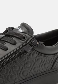 Zapatilla negra con cuero liso y un lateral texturizado que presenta un patrón de logotipos. Incluye cordones negros y un cierre de cremallera en el lateral.