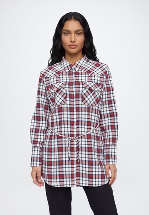 Une femme porte une longue chemise à carreaux rouges, bleu marine et blancs, avec un lien à nouer à la taille et deux poches poitrine, assortie à un pantalon noir.