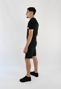 Conjunto atlético para hombre que incluye una camiseta negra de manga corta con un reverso verde oscuro, shorts verdes a juego y zapatillas negras.