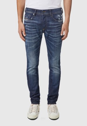 Homme portant un jean slim bleu foncé délavé avec légère usure et des baskets blanches, debout devant un fond uni.