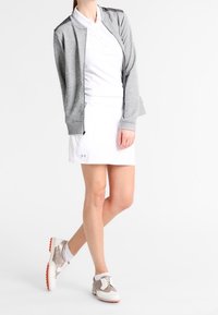 Giacca zip grigia chiara, polo bianca, gonna bianca con logo e sneaker beige con dettagli arancioni. Texture lisce e design sportivo.