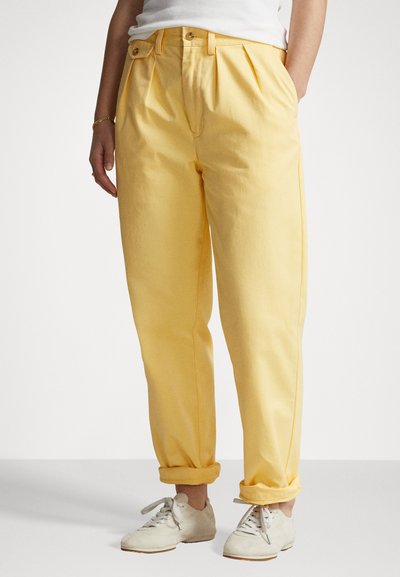 Pantaloni plisați galbeni cu o croială lejeră, manșete răsucite, două butoane la talie și buzunare laterale. Asortați cu adidași albi.