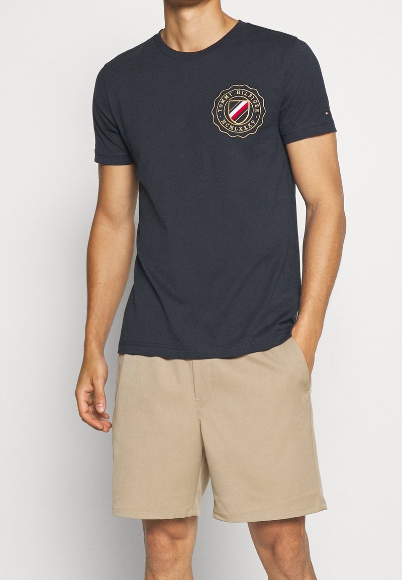 T-shirt en coton bleu marine, col rond, manches courtes, avec un logo circulaire aux accents rouges et or sur la partie gauche de la poitrine, porté avec un short beige.