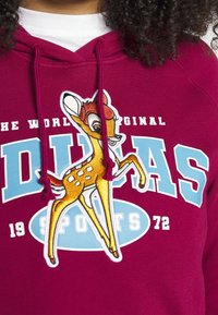 Child wearing a burgundy hoodie with blue and white text and a colorful embroidered cartoon fawn patch on the front.