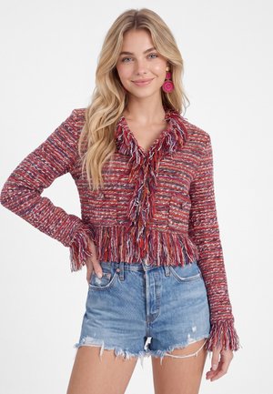 Vrouw met lang blond haar die een rode gebreide trui met franjes en blauwe gescheurde spijkershort draagt, glimlachend met één hand op haar heup.