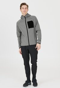 Graue Zip-Hoodie mit einer schwarzen Brusttasche, kombiniert mit schwarzen Hosen und schwarzen Sportschuhen. Glatte Textur und figurbetontes Design.