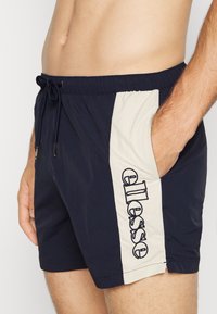 Bañadores de hombre en azul marino con un panel lateral beige que presenta el logotipo bordado de "ellesse", cinturilla elástica y bolsillos laterales.