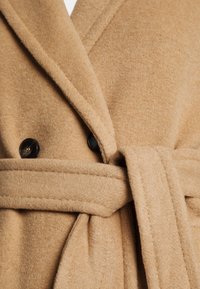 Gros plan d'un manteau en laine brun clair avec de larges revers, des boutons noirs et une ceinture en tissu nouée à la taille.