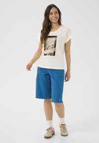 T-shirt couleur crème avec un graphisme sur le thème de la nature en noir et or, associé à un short en denim de longueur genou. Des baskets et des chaussettes blanches complètent la tenue.