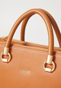 Borsa in pelle beige con hardware dorato, doppi manici, cuciture bianche e chiusura con zip. Presenta il nome del brand impresso sul davanti.