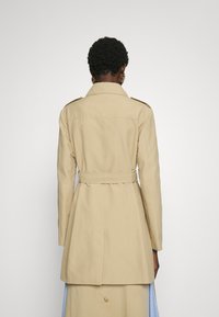 Trench-coat beige avec une taille ceinturée, des épaulettes, des manches longues et une fente à l'arrière. Tissu lisse avec une coupe ajustée.