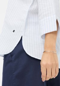 White and light blue striped shirt with short sleeves, curved hem, and side slits. Silver bracelet on a hand, paired with dark navy bottom.