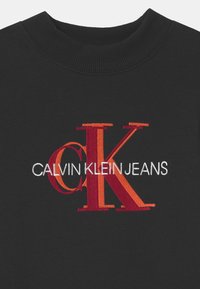 Svart plagg med ribbad krage och broderad röd och orange "cK" logotyp som överlappar vit "CALVIN KLEIN JEANS" text på framsidan.