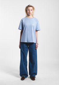 Camiseta de algodón azul claro con mangas cortas y cuello redondeado, que presenta un logo blanco en el lado izquierdo del pecho. Combinada con unos vaqueros de mezclilla azul de corte holgado.