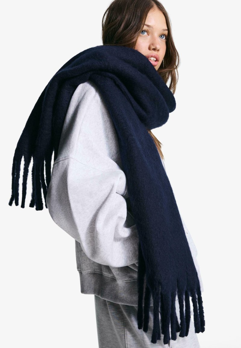 Mørkeblå, strikket tørklæde lavet af blødt stof, med frynser i enderne. Båret afslappet over en lysegrå oversized sweatshirt.