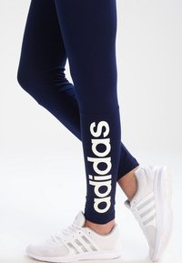 Ben klädda i marinblå Adidas leggings med vitt logotyp på vaden, tillsammans med vita Adidas sneakers.