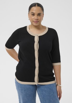 Frau mit dunklem Haar in einem schwarzen, kurzärmeligen, geknöpften Cardigan mit beiger Einfassung, blauen Jeans und silbernen Creolen-Ohrringen und Armband.