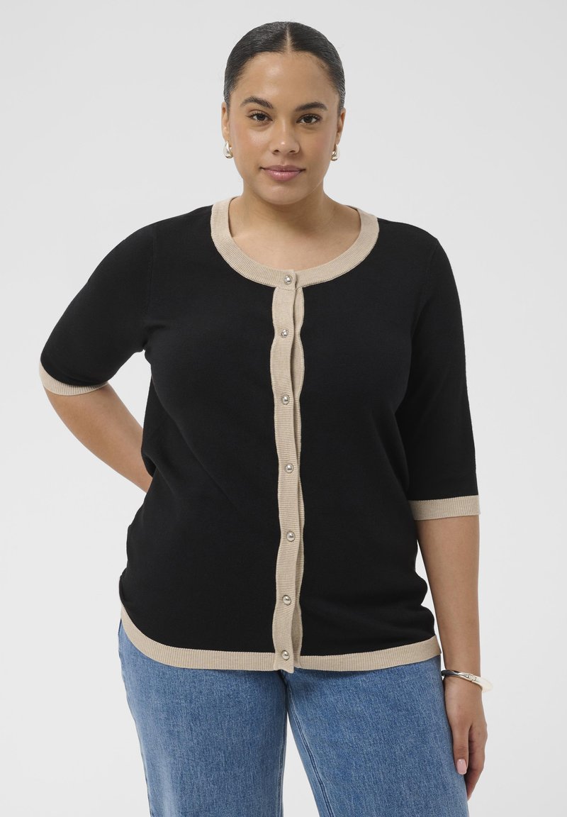 Femme aux cheveux foncés portant un cardigan noir à manches courtes boutonné avec bord beige, jean bleu, boucles d'oreilles créoles et bracelet argentés.