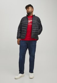 Giubbotto nero imbottito con un design trapuntato, indossato sopra una maglietta grafica rossa e abbinato a jeans blu e sneakers bianche.
